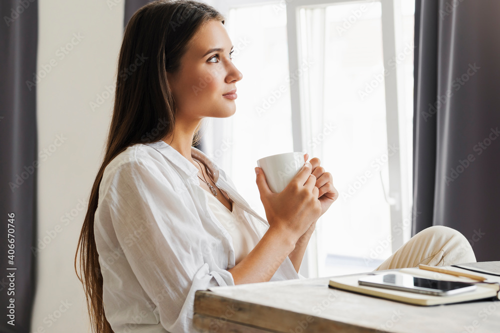 Beautiful brunette girl is sitting at table in front of laptop, holding cup of coffee with her hands. Woman resting at home, enjoying watching movies, online services, pleasant pastime Beautiful brunette girl is sitting at table in front of laptop, holding cup of coffee with her hands. Woman resting at home, enjoying watching movies, online services, pleasant pastime