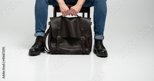 Man in a blue jeans and black boots sits on a chair with a brown men's shoulder leather bag for a documents and laptop on a white floor. Mens leather satchel, messenger bags, handmade briefcase.