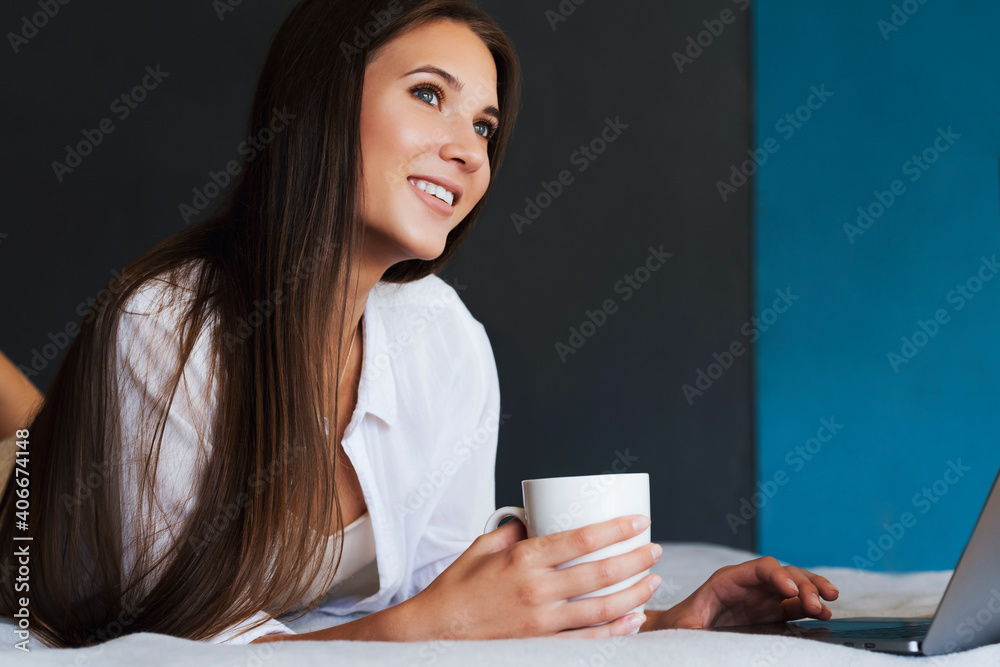 Millennial girl is resting on bed in white shirt, holding cup of coffee in her hand. Beautiful brunette woman uses laptop to watch online content, TV series, online training, work remotely from home Millennial girl is resting on bed in white shirt, holding cup of coffee in her hand. Beautiful brunette woman uses laptop to watch online content, TV series, online training, work remotely from home