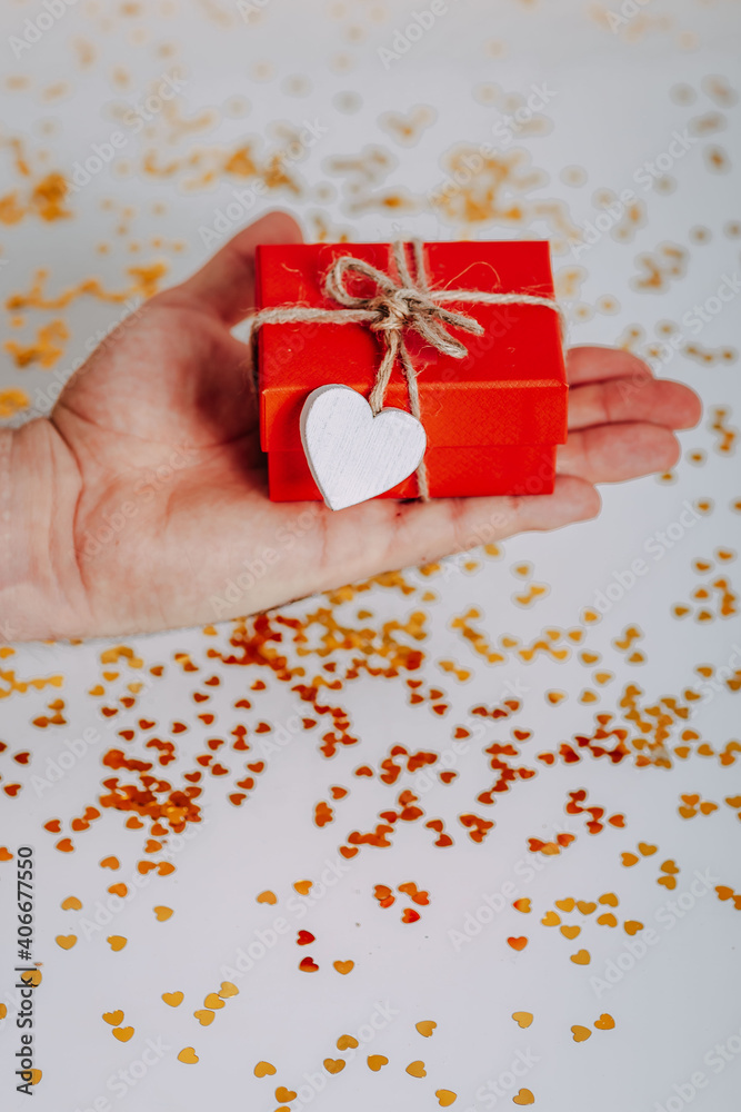 Man heand with small red gift box with golden glitters around. Red present with heart on the rope on white background. Saint Valentine's Day present. Heart-shaped golden glitters.