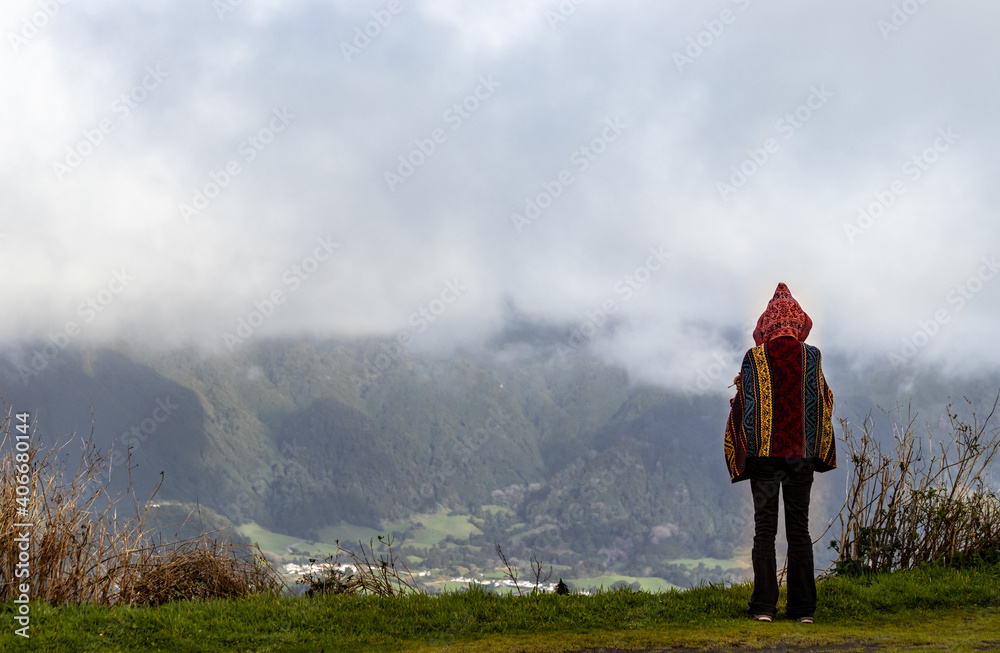 Fototapeta premium Person with hoodie, standing at viewpoint, mountains and clouds, Furnas village, Sao Miguel, Azores islands.