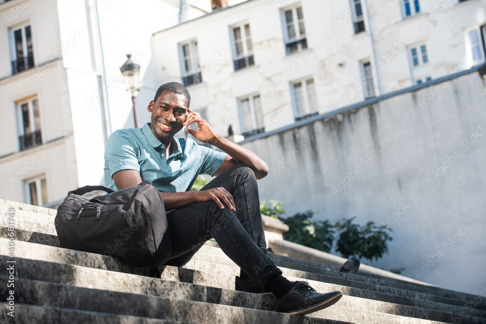 Full body happy young black man sitting on steps in city talking with mobile phone