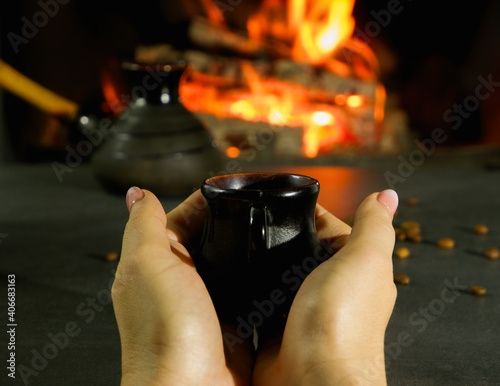A young woman enjoys a coffee brewed in the traditional way in a ceramic dish. Close-up on hands against the background of burning fire and coffee beans.