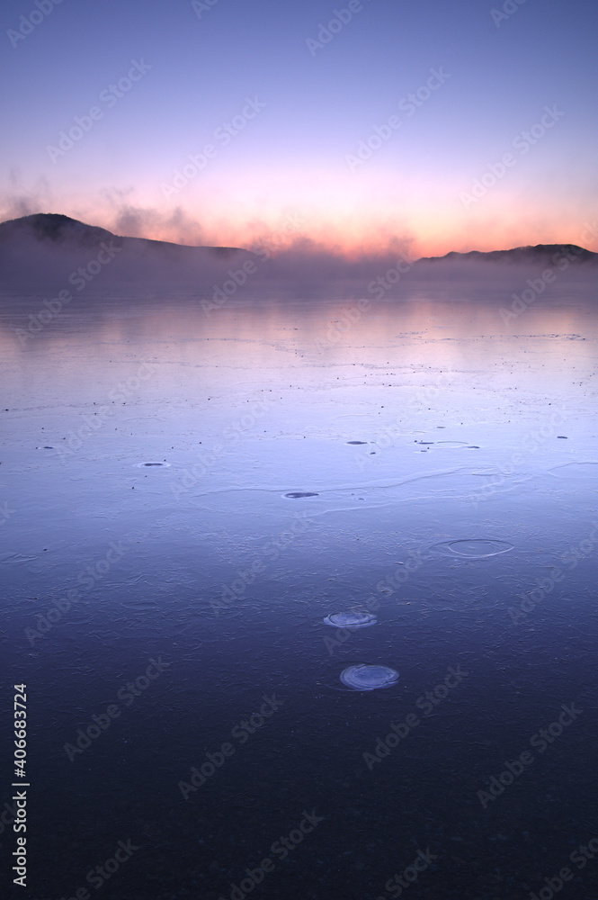 朝靄の漂う夜明けの湖。結氷した湖面に映る空の色。屈斜路湖、北海道、日本。