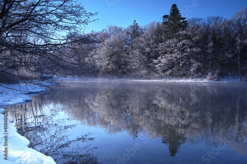 冬の青空の下の湖の風景。湖面に映る湖畔の森。阿寒摩周国立公園の屈斜路湖、北海道、日本。 © Masa Tsuchiya