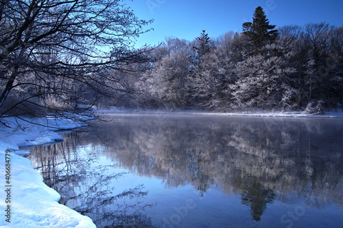 冬の青空の下の湖の風景。湖面に映る湖畔の森。阿寒摩周国立公園の屈斜路湖、北海道、日本。 © Masa Tsuchiya