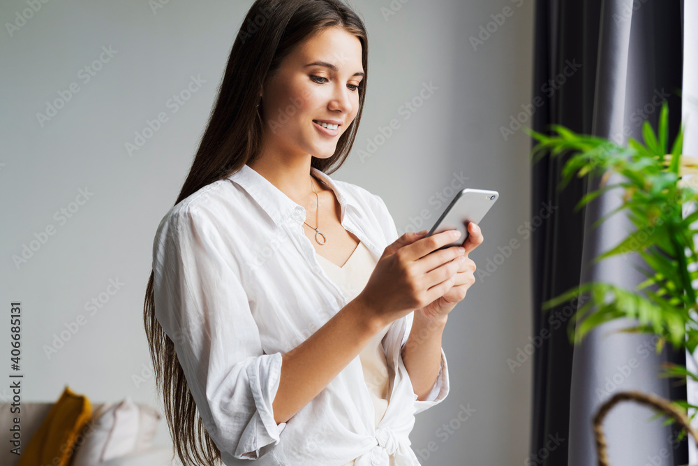 Portrait of business woman, holds smartphone in her hands. Beautiful girl uses mobile phone for work, calls clients, sends text messages. Millennial girl in white shirt stands against white wall Portrait of business woman, holds smartphone in her hands. Beautiful girl uses mobile phone for work, calls clients, sends text messages. Millennial girl in white shirt stands against white wall