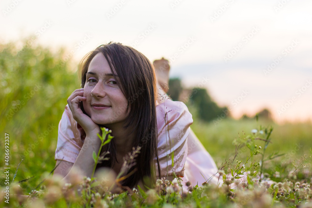 A young beautiful girl in a long pink dress with bare feet lies in a field in the grass and wildflowers holding head on hand and smiles. A young beautiful girl in a long pink dress with bare feet lies in a field in the grass and wildflowers holding head on hand and smiles.