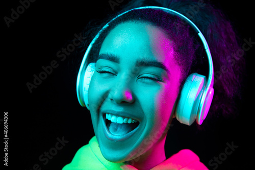 Laughting, close up. African-american woman isolated on dark background in multicolored neon. Listening to music with headphones. Concept of human emotions, facial expression, sales, ad, fashion.