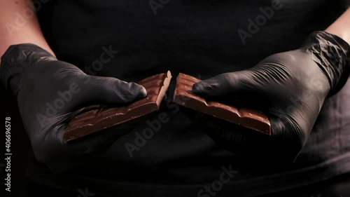 Chef chocolatier in black gloves breaks chocolate bar in half for melting and making sweets, cooking the sweet desserts. Man's hands close up. Slow motion. photo