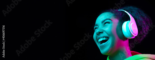 Laughting, flyer. African-american woman isolated on dark background in multicolored neon. Listening to music with headphones. Concept of human emotions, facial expression, sales, ad, fashion.