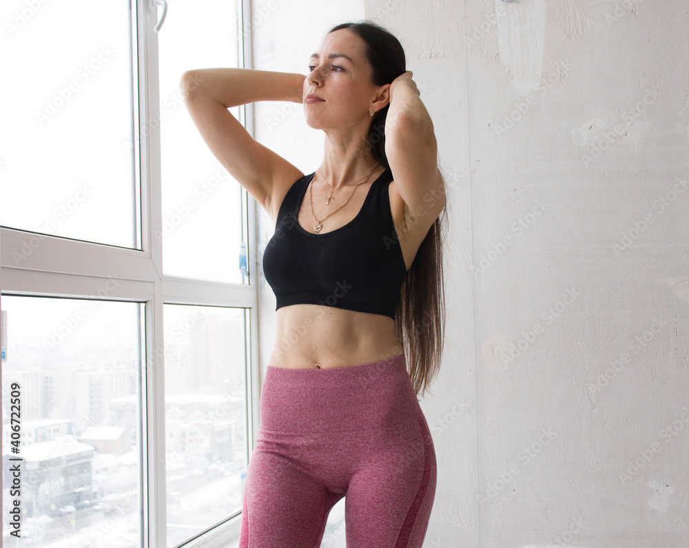 Close-up of a young slender woman before a yoga class, a female instructor after a workout at home or in a club, standing at the window. Healthy lifestyle, hobbies, beauty, well-being concept