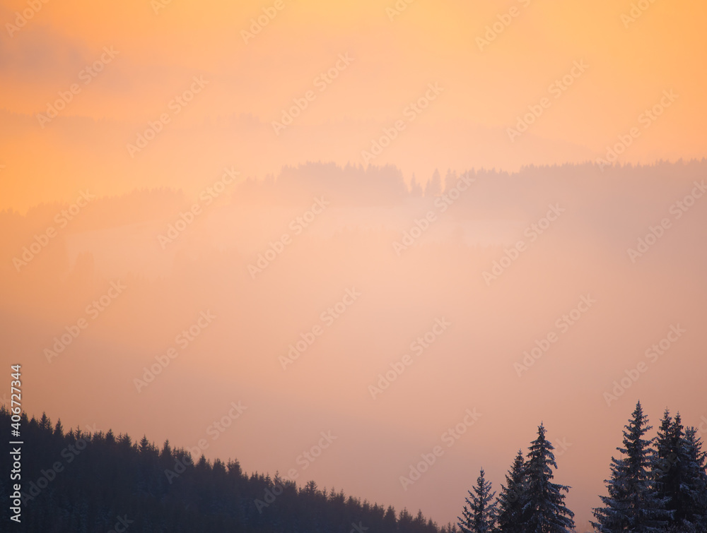 Abstract background of dreamy mauve pink to pastel golden orange fog light over mountain forest silhouettes at winter sunrise or sunset. Magic Valentine moments in foggy mountain valley landscape. 