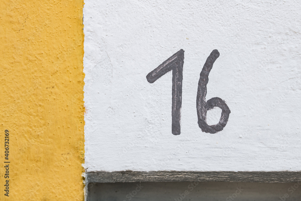 The number 16 roughly hand painted in black on a white wall with a yellow ink strip on the left side of frame. Detail of facade with house number sixteen. Minimalist photograph with straight lines The number 16 roughly hand painted in black on a white wall with a yellow ink strip on the left side of frame. Detail of facade with house number sixteen. Minimalist photograph with straight lines