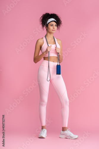 Fototapeta Naklejka Na Ścianę i Meble -  full length of curly young sportswoman standing with jumping rope on pink