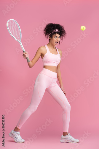 full length of curly woman in sportswear holding racket while playing tennis on pink