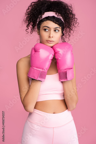 curly young woman in sportswear and boxing gloves looking away isolated on pink