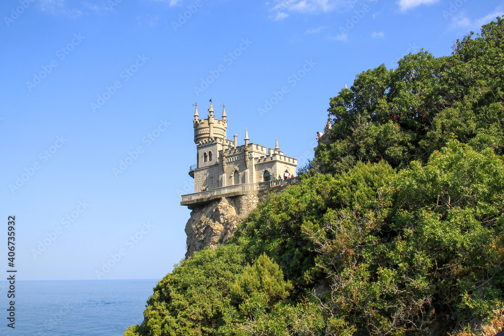 View of the Swallow's nest from the shore on a sunny day. Bright  blue sky on a summer day. A large green bush covers the rock with a castle.