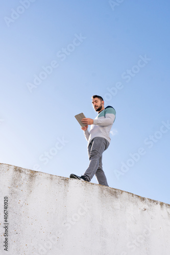 Caucasian boy with a beard sitting on top of a wall with an I pad making a video call in sunlight outdoors and wearing colorful sweater
