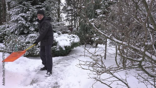 A 61-year-old European clears snow from a path in his garden by the forest. The poodle dog runs happily next to him. Healthy lifestyle concept in old age in winter