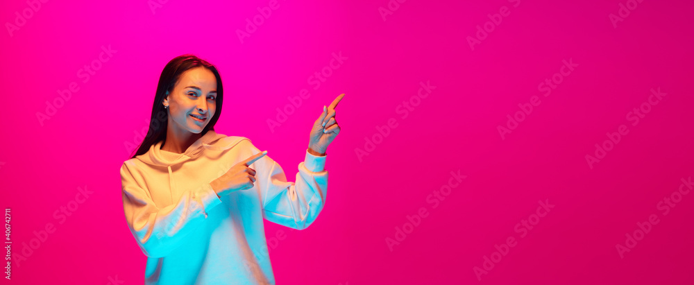 Pointing, flyer. Caucasian woman's portrait isolated on pink studio background in mixed neon light. Beautiful female model. Concept of human emotions, facial expression, sales, ad, fashion. Copyspace. Pointing, flyer. Caucasian woman's portrait isolated on pink studio background in mixed neon light. Beautiful female model. Concept of human emotions, facial expression, sales, ad, fashion. Copyspace.