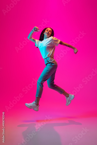 Jumping high, looks cheerful. Caucasian woman's portrait on pink studio background in mixed neon light. Stylish outfit. Concept of human emotions, facial expression, sales, ad, fashion. Copyspace.