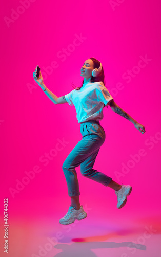 Jumping high. Caucasian woman's portrait on pink studio background in mixed neon light. Listening to music with phone. Concept of human emotions, facial expression, sales, ad, fashion. Copyspace.