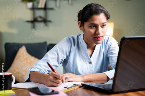 Young business woman writing down notes by looking laptop - concept of employee or student online training class at home. photo