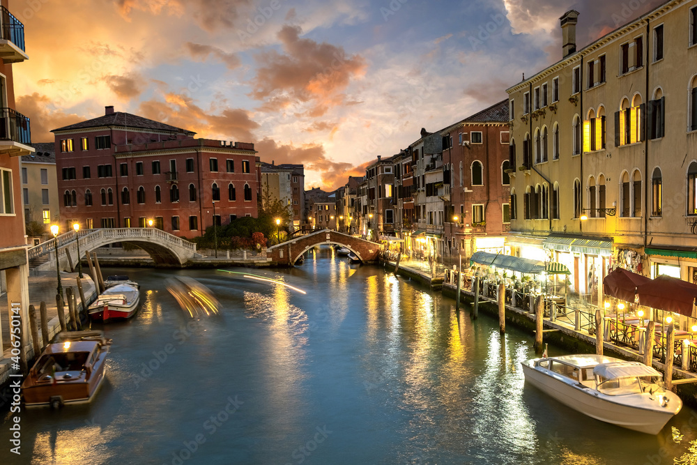 Night view of illuminated old buildings, floating boats and light reflections in canal water in Venice, Italy.