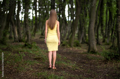 Back view of a slim woman in yellow dress standing in moody dark forest.