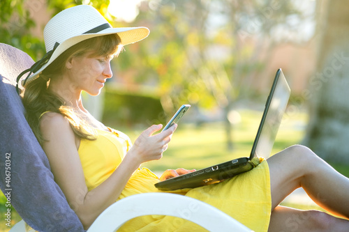 Girl student in yellow summer dress resting on green lawn in summer park studying on computer laptop texting on mobile cell phone. Doing business and learning during quarantine concept.