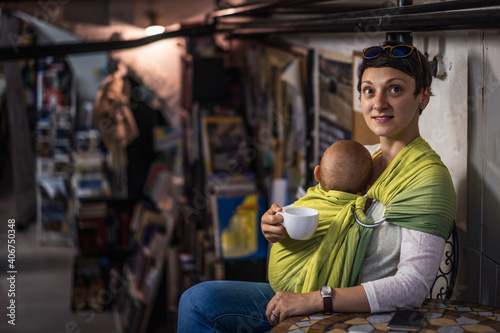 A young mother with her baby in a carrier sling drinking coffee. Beautiful happy young mother with baby carrier. 