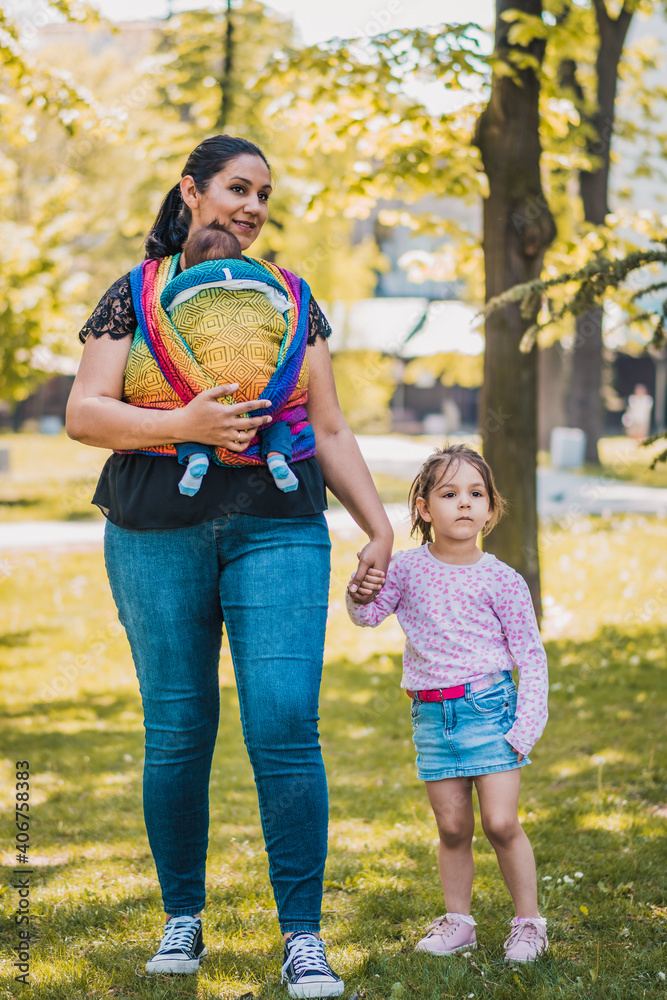 Fototapeta premium Beautiful happy young mother with baby and child carrier in park. Mom walking with infant in baby carrier. Happy mother carrying her child by ergonomic baby carrier.