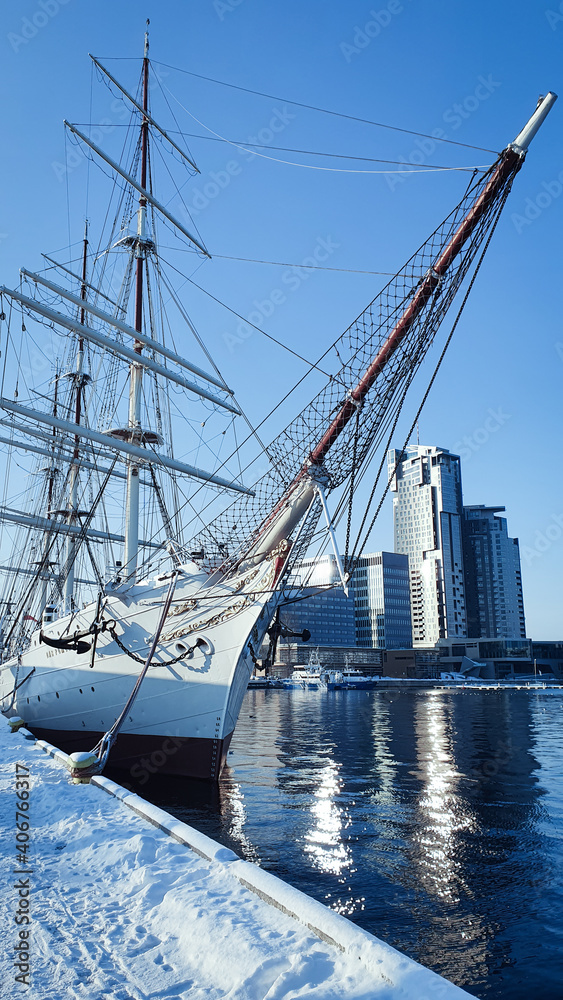 Sailing ship at the snow-covered quay in the Polish city of Gdynia. A skyscraper in the background. A photo in a silvery color. Sailing ship at the snow-covered quay in the Polish city of Gdynia. A skyscraper in the background. A photo in a silvery color.