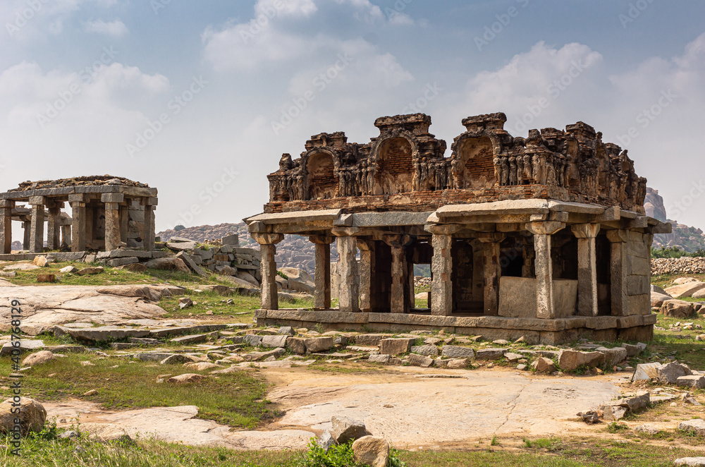 Hampi, Karnataka, India - November 5, 2013: Ancient Temple. Most conserved red stone ruin with ceiling and empty niches under blue cloudscape on rocky plateau with grass up front.