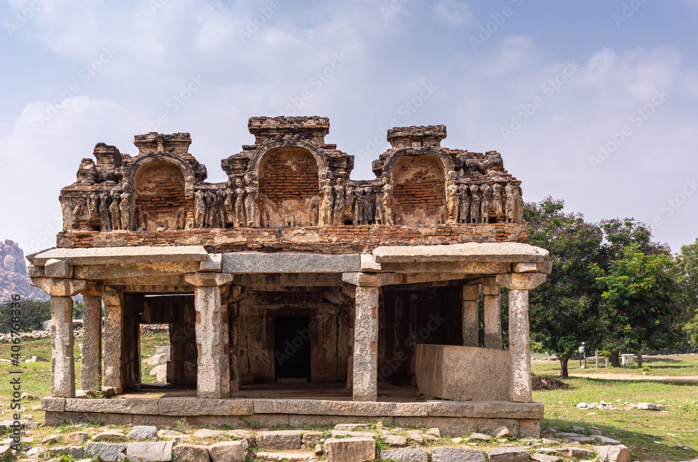 Hampi, Karnataka, India - November 5, 2013: Ancient Temple. Closeup of beige stone sculpted ruinous sanctuary with red empty niches on top under blue cloudscape and some green foliage.