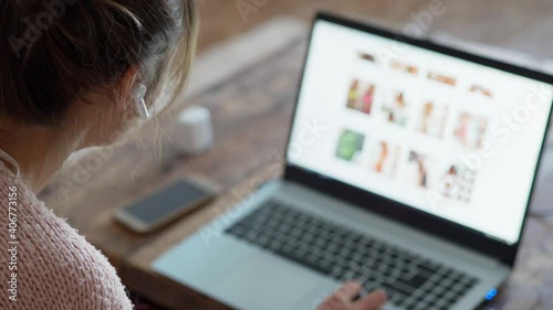 young unrecognizable woman with wireless earphones sits at wooden desk at home, browsing site of online store on laptop and making online order from home by phone. focus on earphone