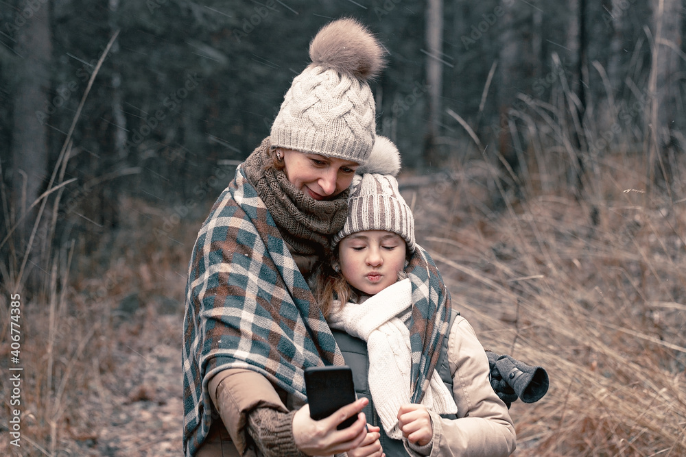 Mom and daughter are taking a selfie. Autumn forest background. Winter clothes fashion