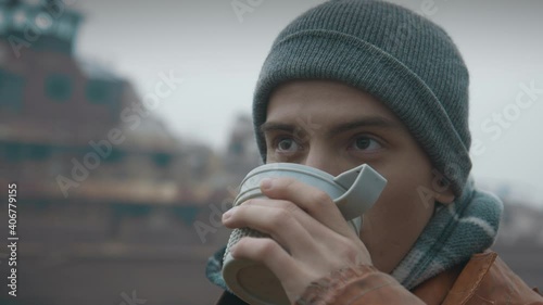 Close-up, portrait: a young male traveler drinking hot tea against the background of an abandoned old rusty ship, looking away. The guy is alone, drinking coffee, social isolation, decay. Slow mo, 4K