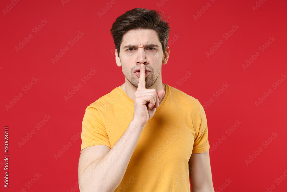 Young unshaved caucasian handsome man 20s wearing casual basic yellow t-shirt saying hush be quiet with finger on lips shhh gesture keeping secret isolated on red color background studio portrait. Young unshaved caucasian handsome man 20s wearing casual basic yellow t-shirt saying hush be quiet with finger on lips shhh gesture keeping secret isolated on red color background studio portrait.