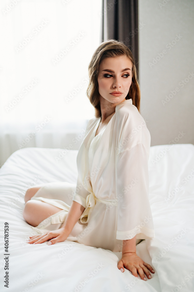 beautiful woman with makeup, long hair sits in white robe on bed beautiful woman with makeup, long hair sits in white robe on bed