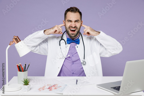 Irriatated nervous male doctor man wearing white medical gown suit sit at desk work on computer in clinic office cover ears with fingers shout scream isolated on violet background studio portrait