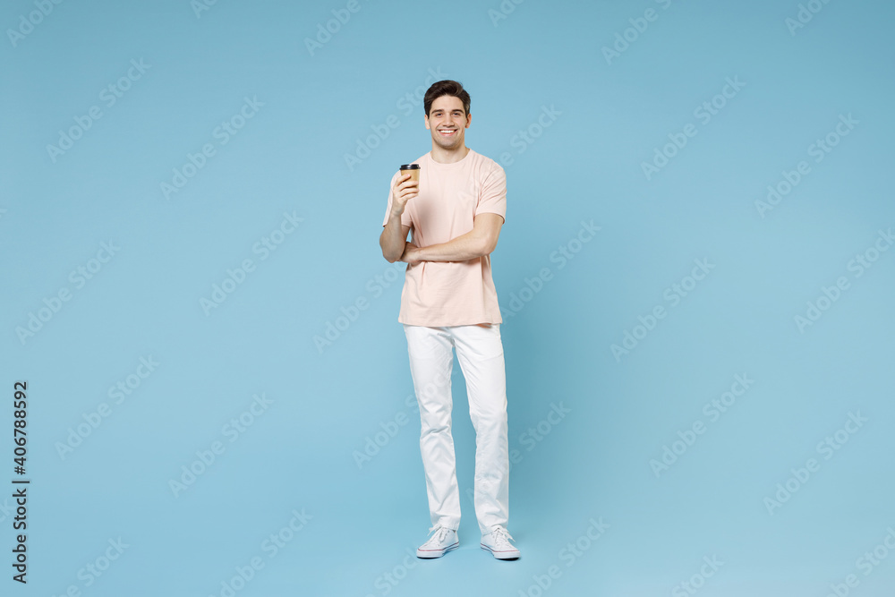 Full length of young caucasian attractive happy smiling student man 20s in beige t-shirt white pants hold paper cup of hot coffee drink look camera isolated on blue color background studio portrait. Full length of young caucasian attractive happy smiling student man 20s in beige t-shirt white pants hold paper cup of hot coffee drink look camera isolated on blue color background studio portrait.