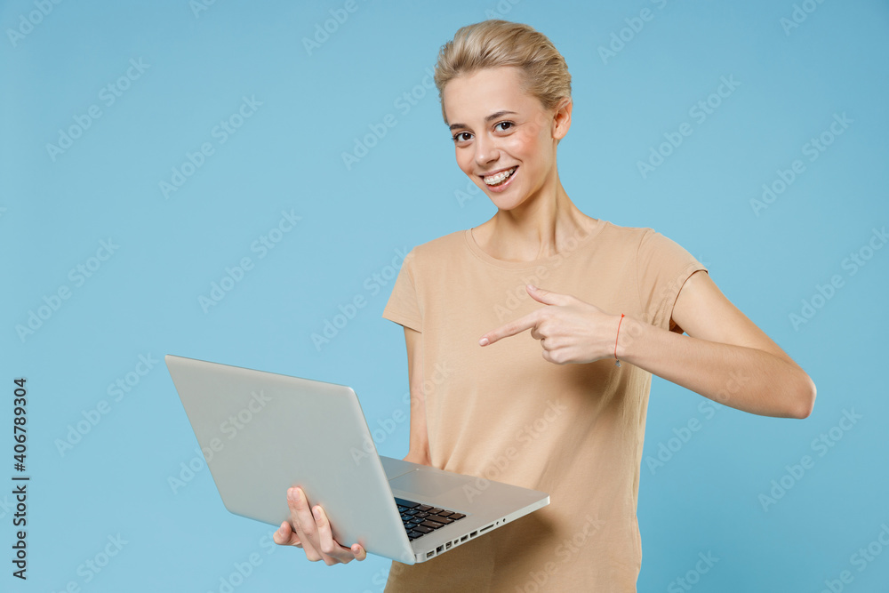 Young smiling caucasian blonde woman 20s with short haircut dental braces in casual beige t-shirt chatting point index finger on modern laptop pc computer isolated on blue background studio portrait. Young smiling caucasian blonde woman 20s with short haircut dental braces in casual beige t-shirt chatting point index finger on modern laptop pc computer isolated on blue background studio portrait.