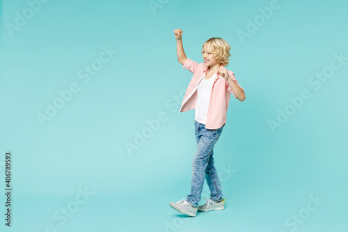 Full length side view of funny little curly kid boy 10s years old in pink shirt clenching fists like winner isolated on blue turquoise background children studio portrait. Childhood lifestyle concept.