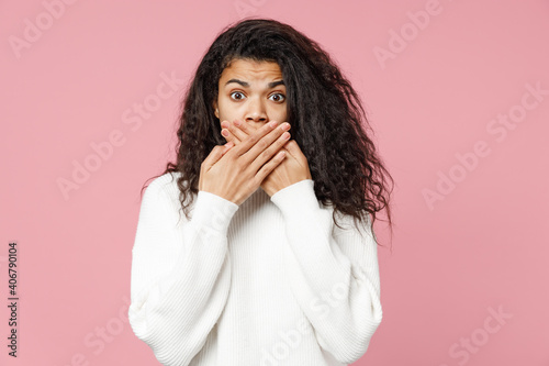 Young shocked scared frightened wonder african american woman 20s curly hair in white knitted sweater cover mouth with palms hands isolated on pastel pink background studio portrait Lifestyle concept
