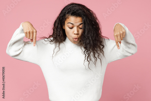 Young african american shock woman 20s curly hair in white casaul knitted sweater pointing index fingers down on copy space workspace mock up area isolated on pastel pink background studio portrait photo