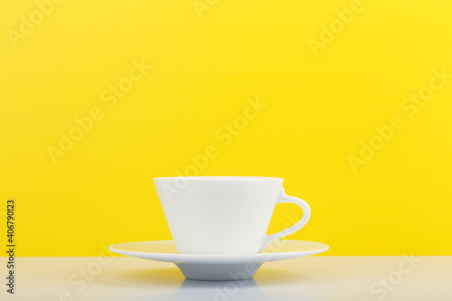 Minimalistic still life with white glossy cup of coffee on white table against yellow background with copy space. 