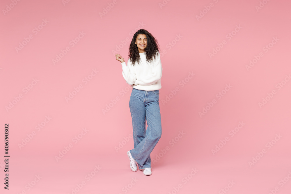 Full length young smiling cute african american woman 20s curly hair wear  white casual knitted sweater jeans look camera hold hands crossed folded  isolated on pastel pink background studio portrait Stock Photo