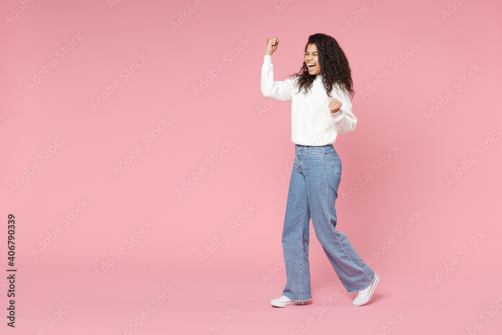 Full length young excited happy overjoyed african american woman 20s curly hair in white knitted sweater jeans do winner gesture clench fist isolated on pastel pink color background studio portrait Full length young excited happy overjoyed african american woman 20s curly hair in white knitted sweater jeans do winner gesture clench fist isolated on pastel pink color background studio portrait